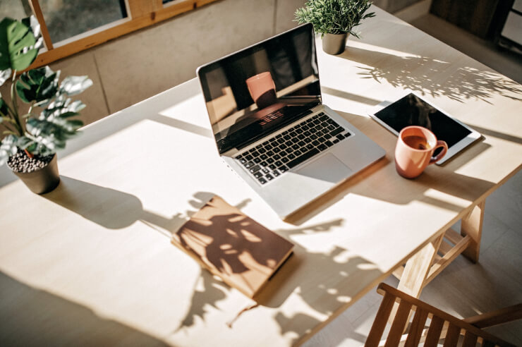 Wide wooden desk with laptop, against window with sun shining through