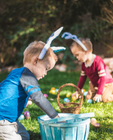 Two children wearing rabbit ears sitting in green field collecting colourful eggs