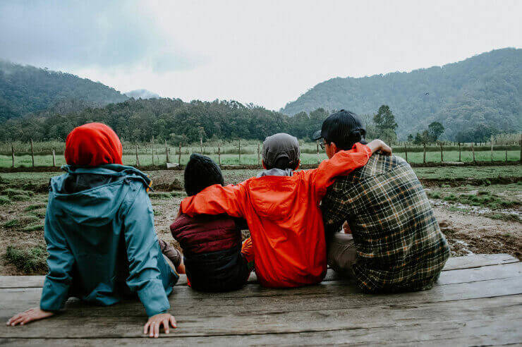Family pictured from behind, sitting on a ledge, looking over a green field with trees