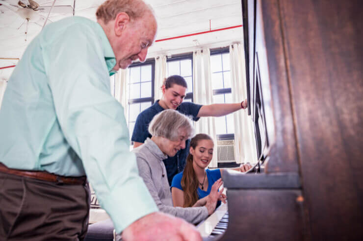 Family gathered around piano, an older lady sits playing with others standing around her