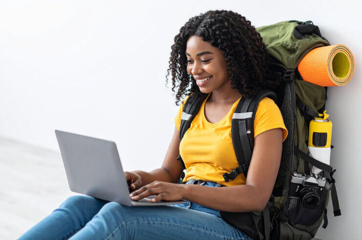 Woman wearing a travel backpack sitting against a wall with her laptop 