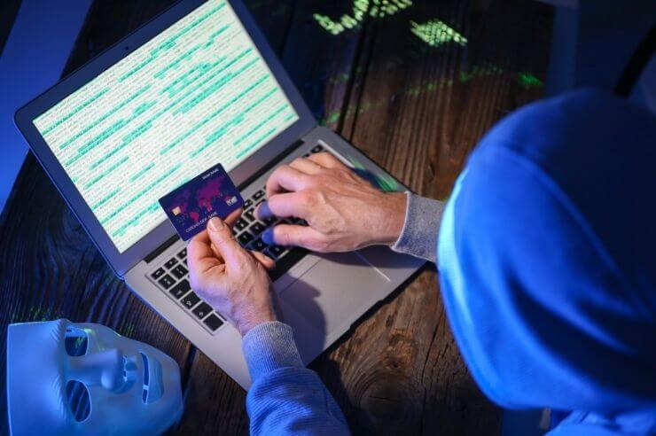 Hacker sitting at laptop inputting details from a credit card, a white mask rests next to him