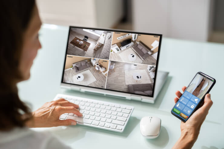 Woman at desk monitoring her home via camera on laptop and smart phone
