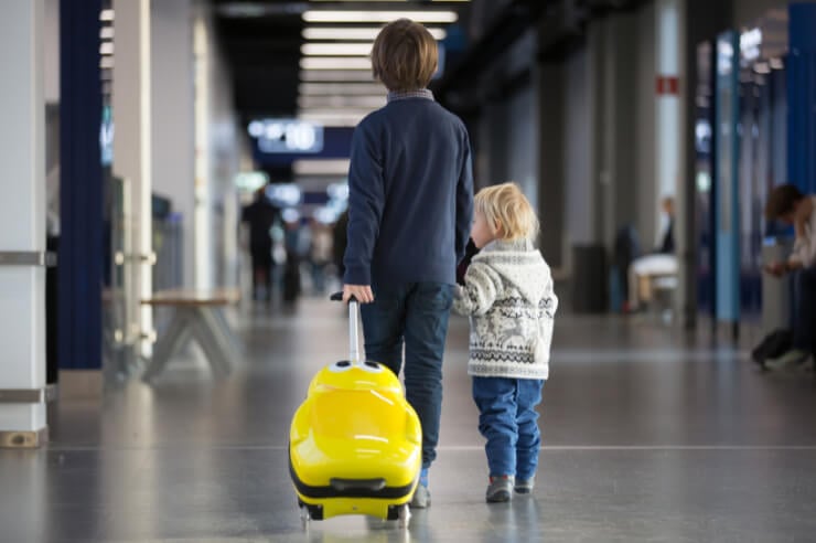 Two children in the airport, one is taller rolling a suitcase, holding hands with the shorter child