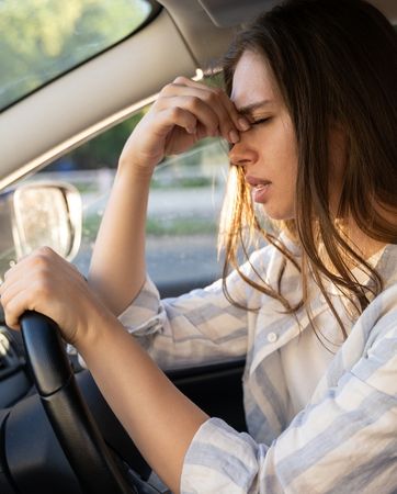 Woman sits at the steering wheel of a car, she looks upset and is pinching the bridge of her nose