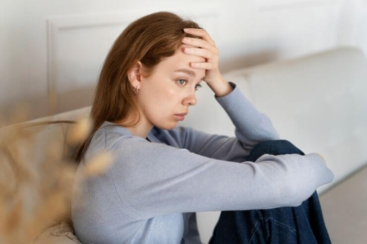 Woman sitting on grey sofa, her knees at her chest, resting her hand on her forehead