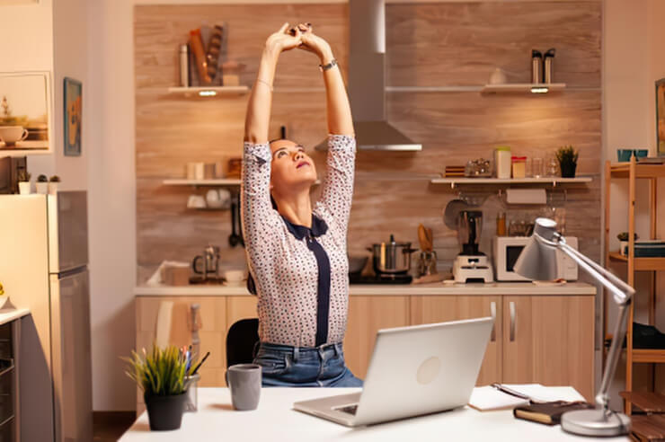 Woman stretching with her hands above her head, her laptop sits open on desk in front of her