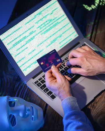 Hacker sitting at laptop inputting details from a credit card, a white mask rests next to him