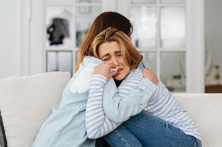 Two women sitting on cream sofa, one appears to be crying while the other comforts her