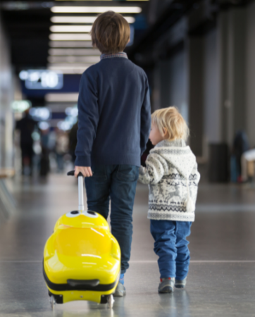 Two children in the airport, one is taller rolling a suitcase, holding hands with the shorter child