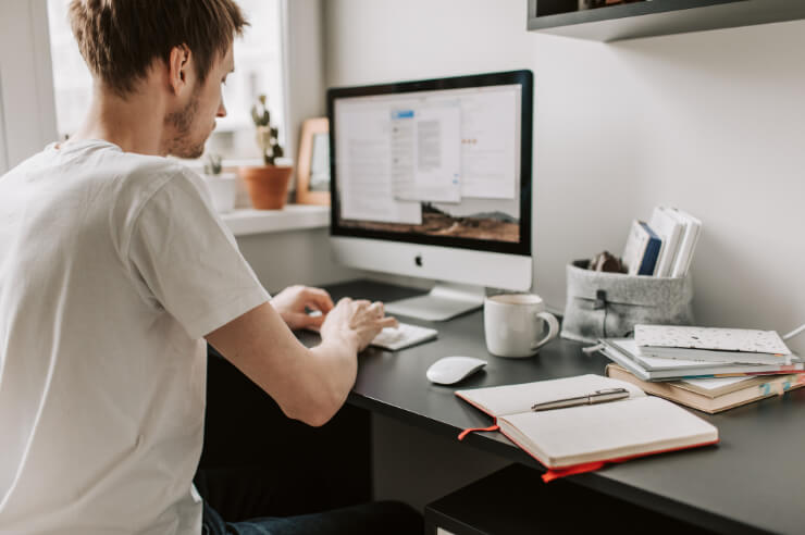 Man working desktop computer at a dark wooden desk. He has a notebook open with a pen resting in it