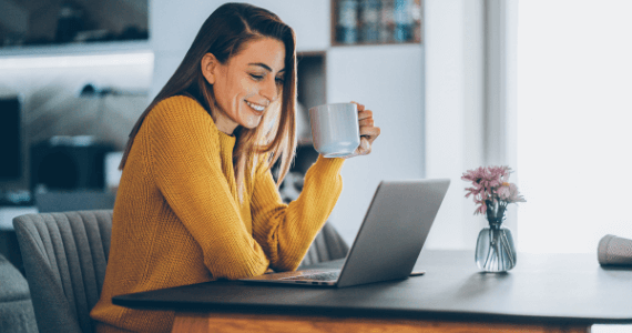 Woman sitting at a table at home, smiling while using a laptop and holding a mug, suggesting a relaxed and productive moment.