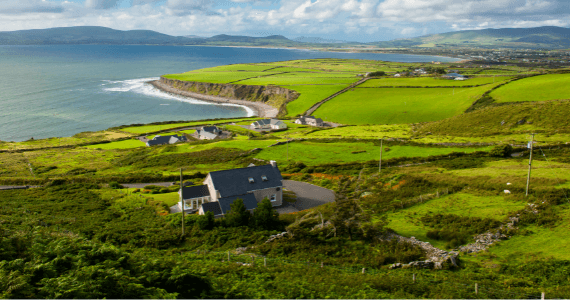 Scenic view of the Irish countryside with green fields, scattered houses, and a dramatic coastline overlooking the ocean under a partly cloudy sky.
