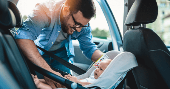 A smiling man securing a baby into a car seat inside a vehicle.