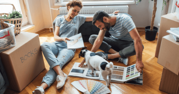 Couple sitting on the floor surrounded by moving boxes, planning home decor with paint samples and their dog.