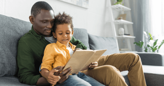 Father and young son sitting on a sofa reading a book together, enjoying a quiet and bonding moment at home.