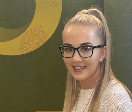 smiling woman with long blonde hair and glasses, wearing a white knit sweater, seated in front of a green background with a yellow circular design, An Post Insurance staff member.