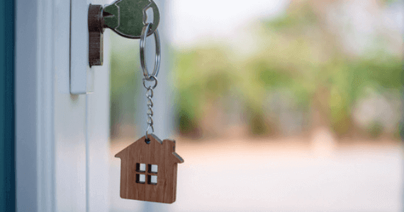 Close-up of a house key in a door lock with a wooden house-shaped keychain.