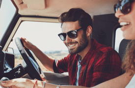 Smiling man in sunglasses driving a vehicle, with a passenger beside him enjoying the ride on a sunny day.
