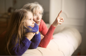 Elderly woman and young girl looking out a window, with the woman pointing at something outside.
