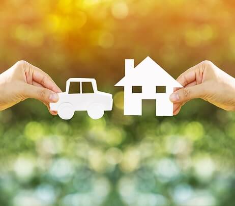 Two hands holding paper cutouts of a car and a house against a blurred natural background with warm sunlight.