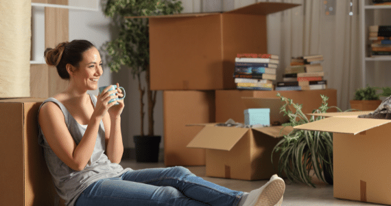 Young woman sitting on the floor with a mug, surrounded by moving boxes and books.