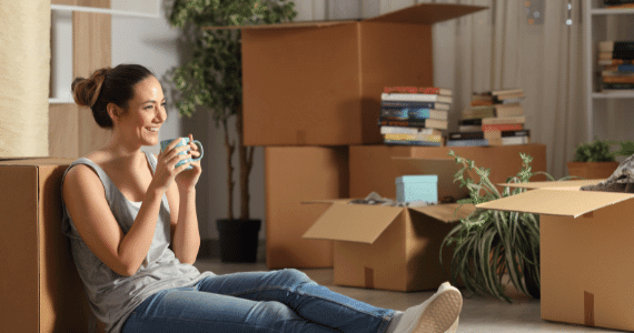 Young woman sitting on the floor with a mug, surrounded by moving boxes and books.
